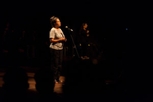 Nayse López, diretora artística e curadora do Festival Panorama, está em pé no centro do palco em frente a um microfone. Está escuro no seu entorno. Ela faz um breve discurso de abertura da noite.