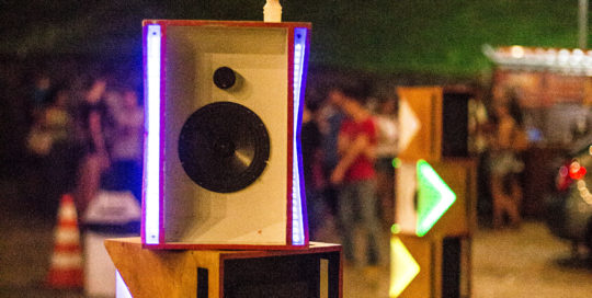 Imagem mostra caixas de som empilhadas. Público aparece desfocado ao fundo. As caixas de som estão iluminadas.