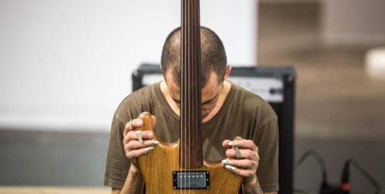 Alejandro Ahmed está no centro, de cabeça baixa, segurando uma guitarra à frente com as duas mãos.