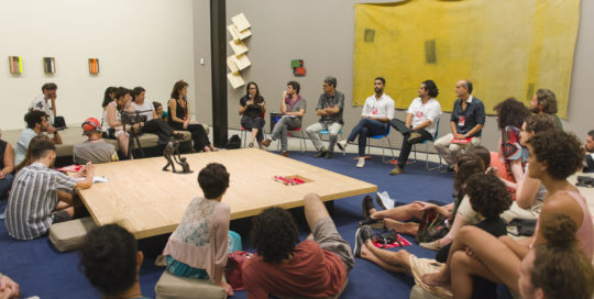 Imagem mostra pessoas sentadas reunidas em torno de uma grande mesa quadrada.